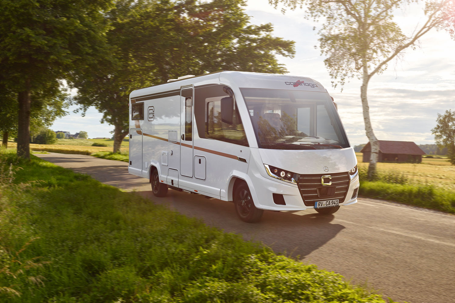 Ein weißes Wohnmobil fährt bei Sonnenschein auf einer Landstraße durch grüne Natur