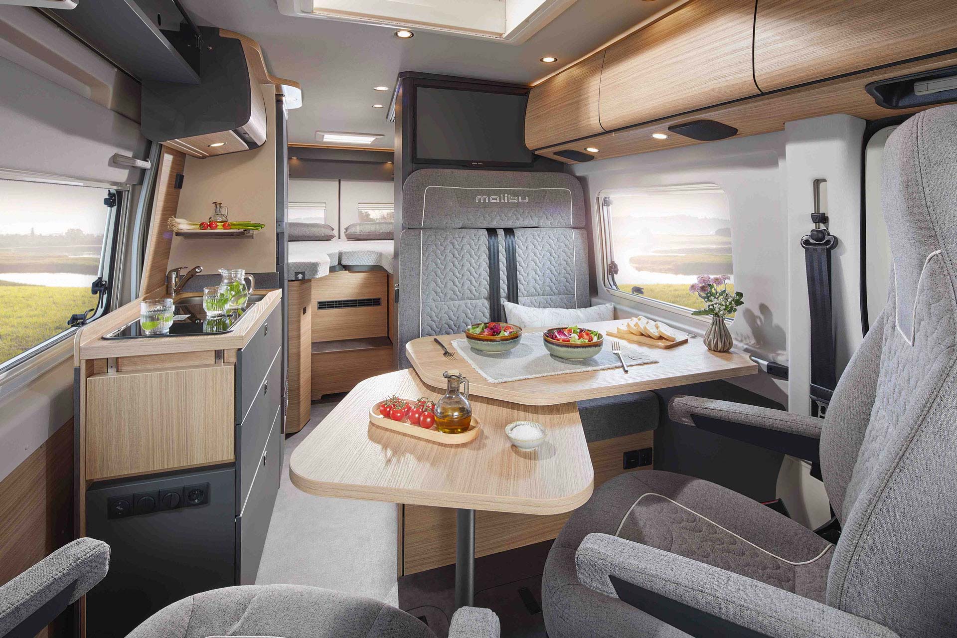 Modern Wohnmobilküche und Essbereich mit hellem Holz, grauen Sitzen und frischem Salat auf dem Tisch