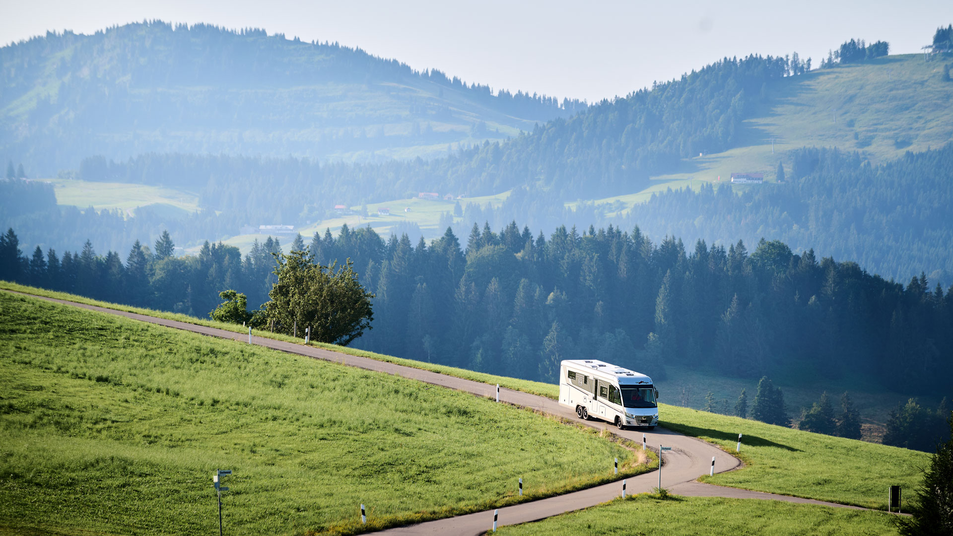 Ein weißes Wohnmobil fährt auf einer kurvigen Straße durch grüne Hügel im Sommer