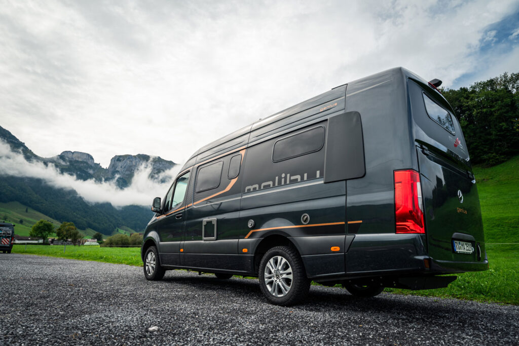 Grauer Campingbus mit orangenen Streifen steht vor grüner Berglandschaft im Nebel