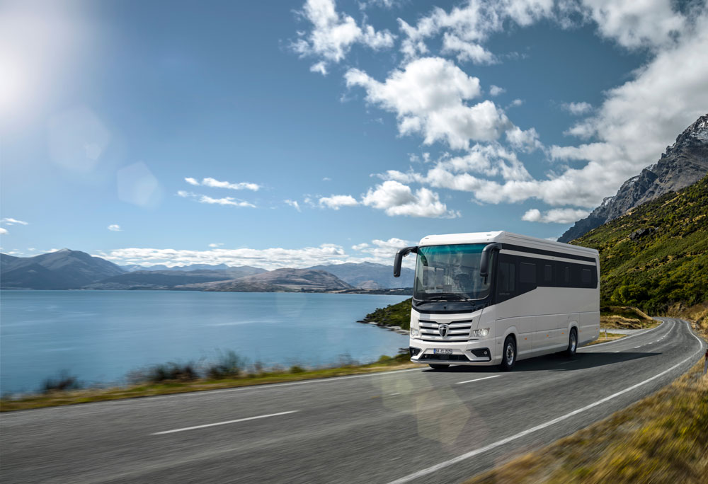 Ein weißer Reisebus fährt bei Sonnenschein auf einer Küstenstraße am Meer entlang