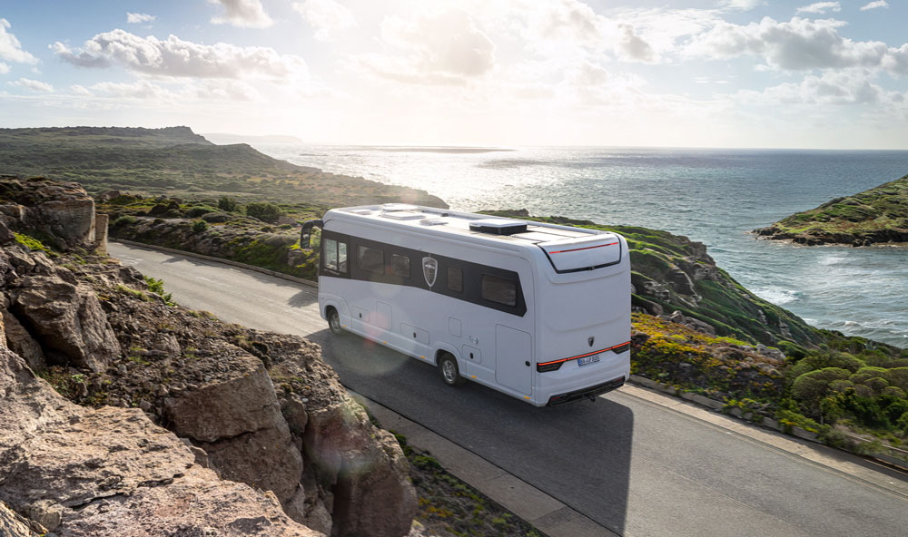 Ein weißes Wohnmobil fährt lächelnd eine Küstenstraße entlang und Sonne spiegelt auf dem Meer