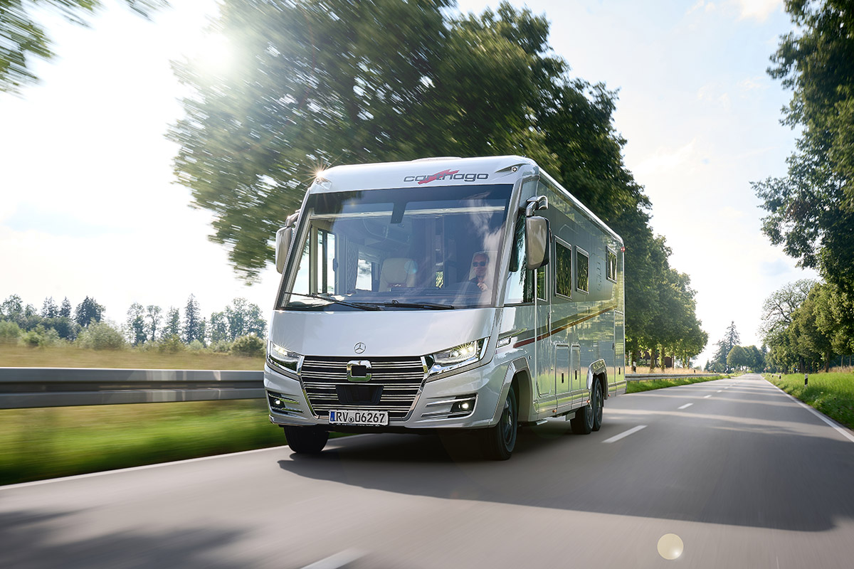 Ein silberfarbener Wohnwagen fährt auf einer sonnigen Landstraße mit Bäumen