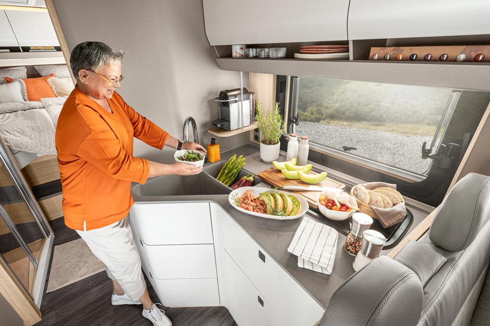 Eine ältere Frau mit grauen Haaren lächelt freundlich in einer modernen Wohnmobilküche und bereitet eine bunte Mahlzeit mit frischen Zutaten zu