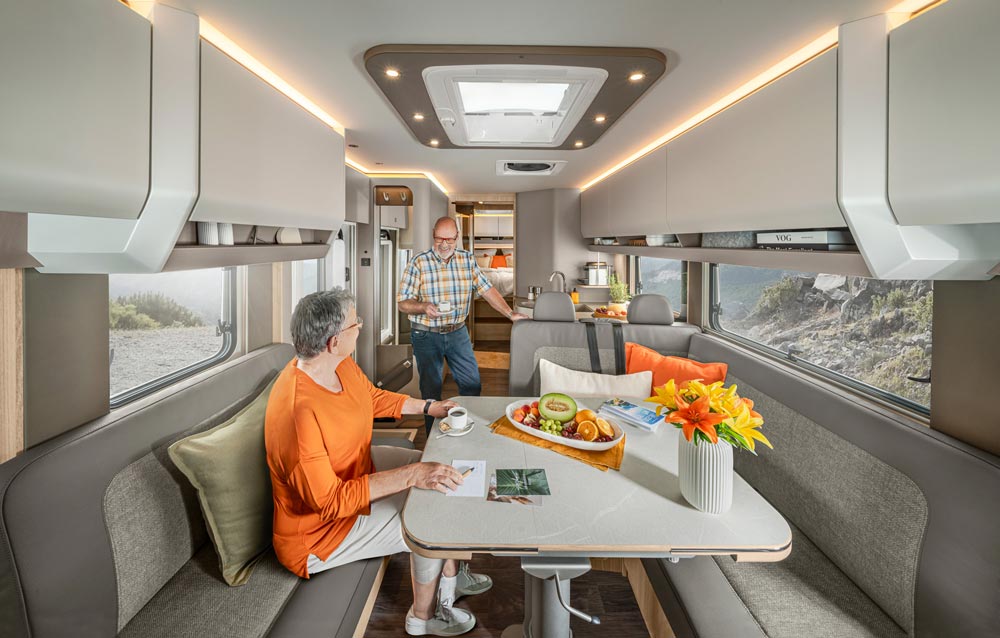 Eine ältere Frau in Orange sitzt lächelnd im hellen Wohnmobil, ein Mann kommt dazu