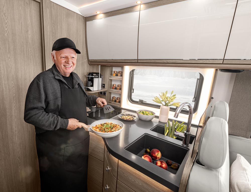 Ein älterer Mann mit schwarzer Kappe lächelt freundlich beim Kochen in einer modernen Camperküche