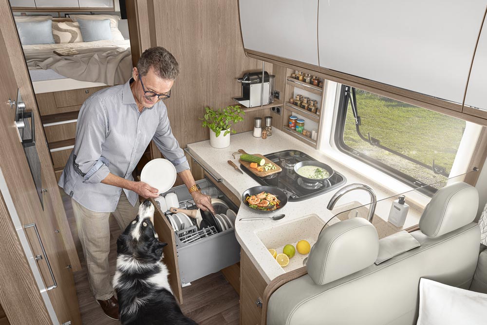 Ein älterer Mann mit Brille lächelt freundlich und füllt die Spülmaschine im Wohnmobil.