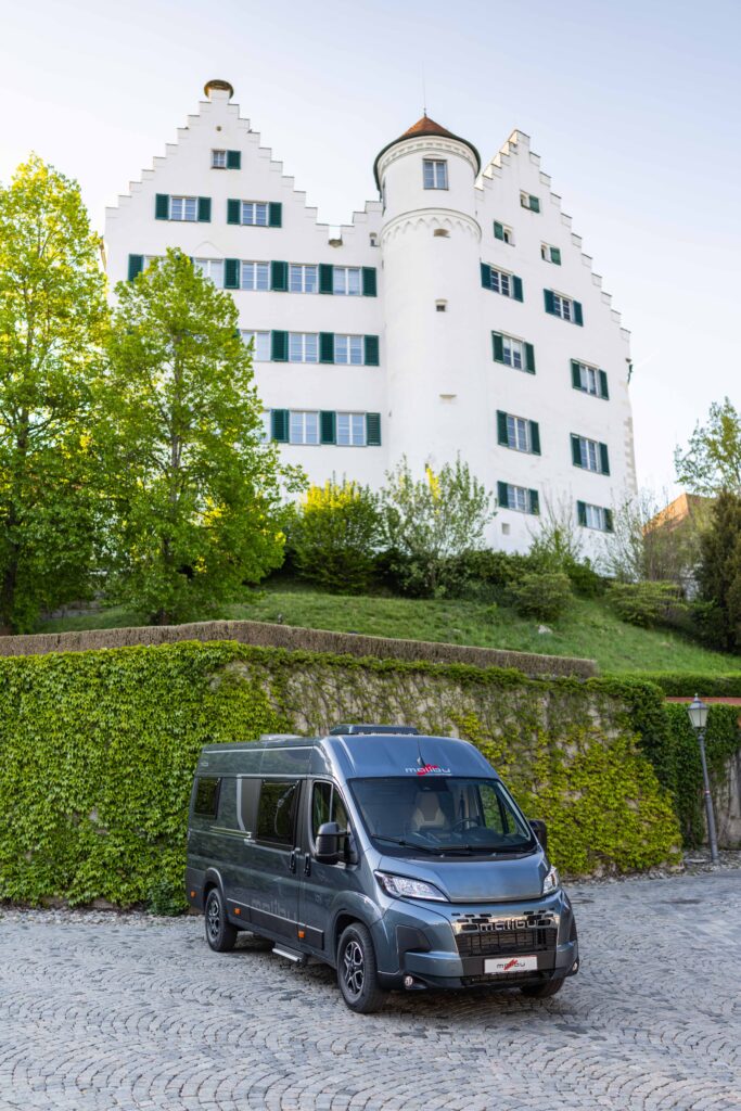Ein grauer Van parkt vor einem weißen Schloss mit Treppengiebeln und rundem Turm