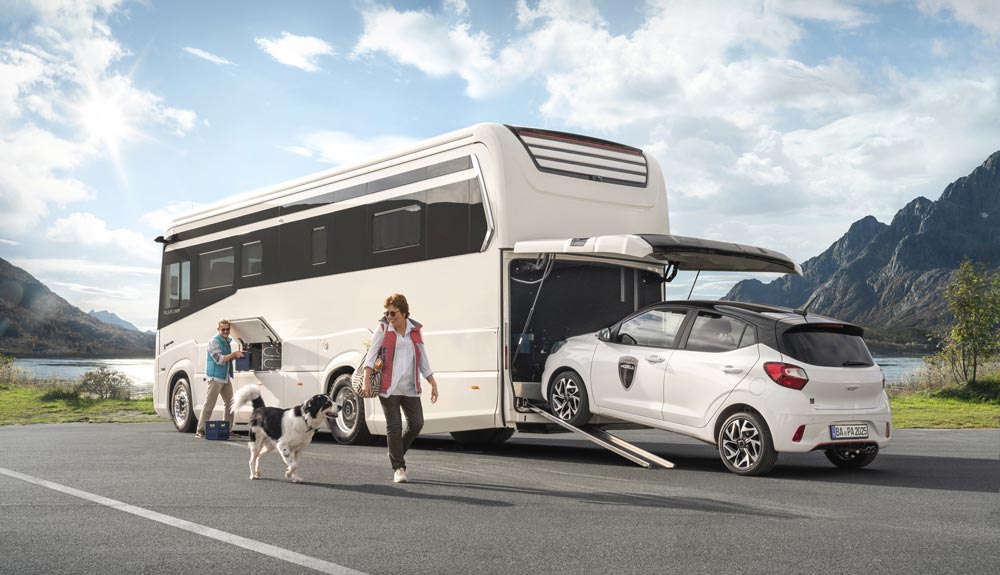 Zwei lächelnde Personen mit Hund stehen neben einem weißen Wohnmobil mit geladenem Auto