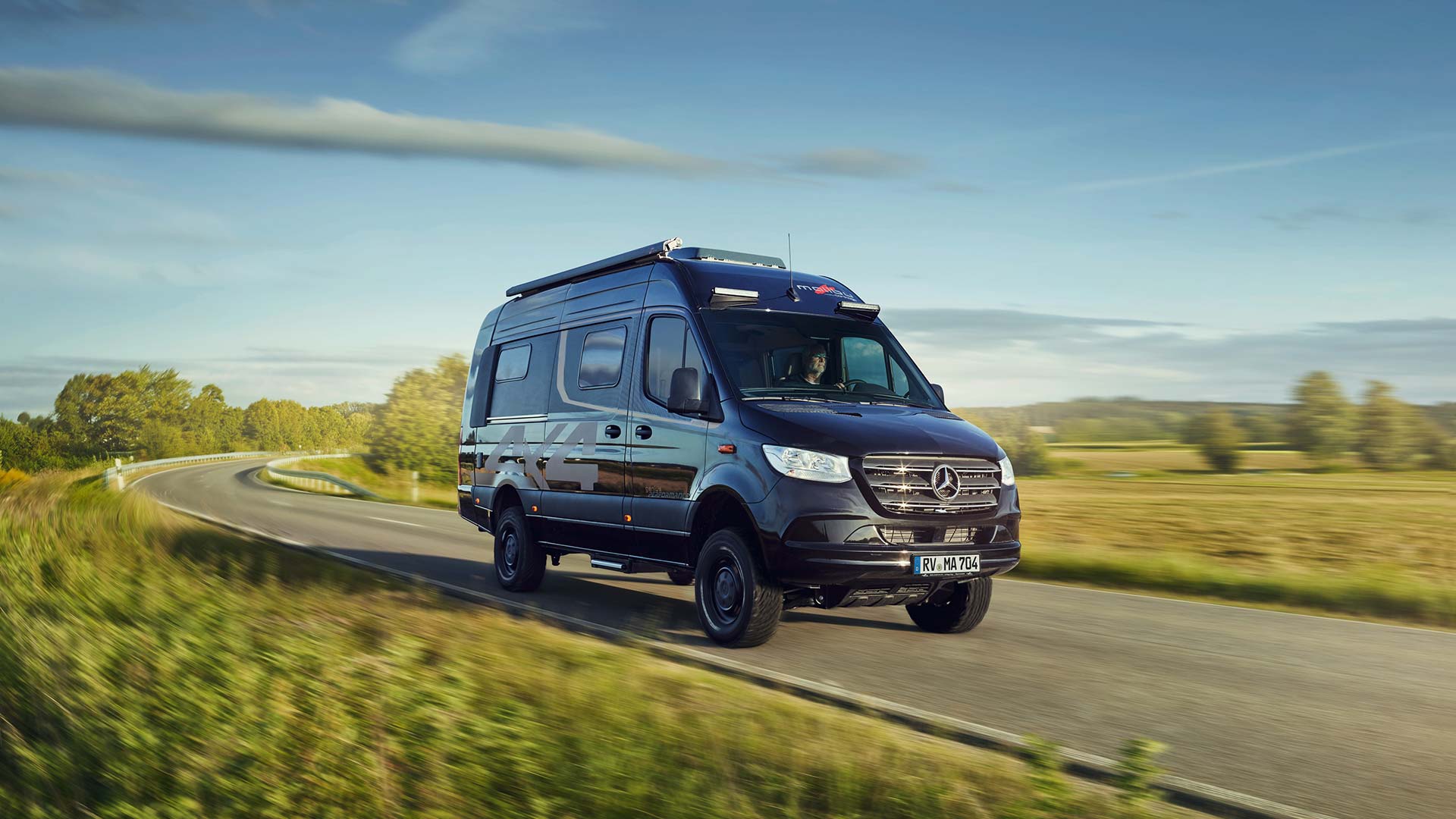 Ein schwarzer Mercedes Sprinter Wohnmobil fährt dynamisch auf einer Landstraße im Sonnenschein