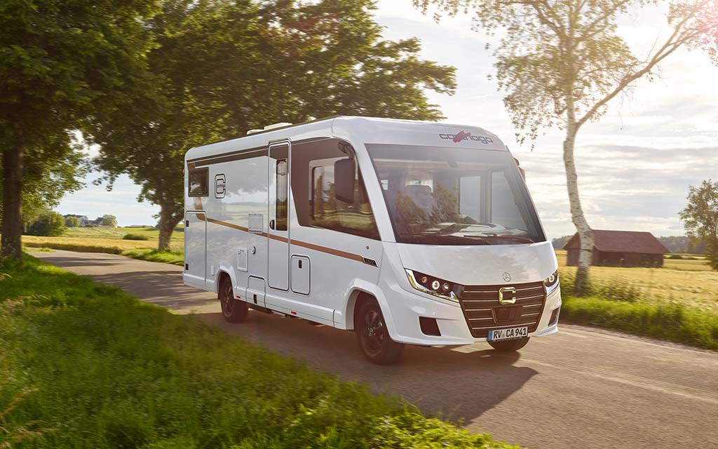 Ein weißes Wohnmobil fährt auf einer Landstraße durch grüne Felder bei Sonnenschein