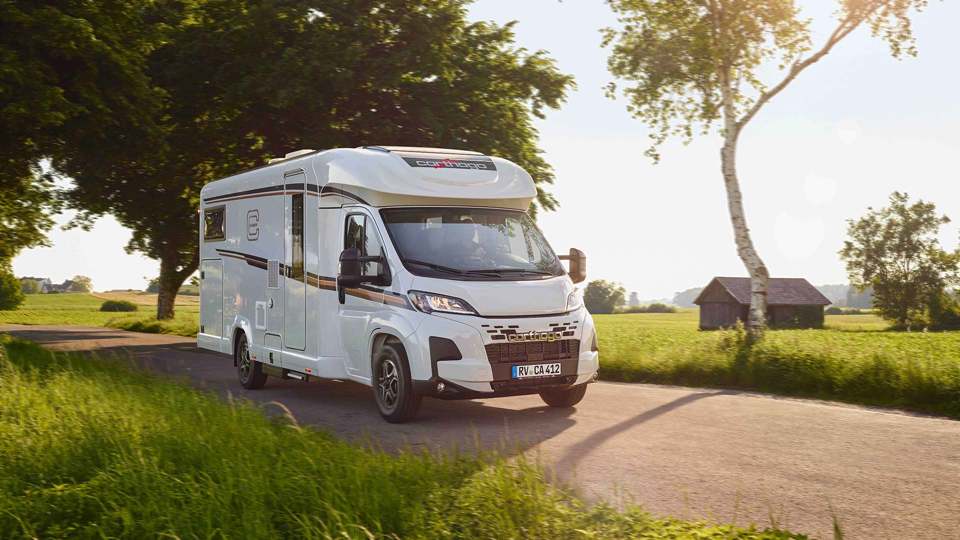 Ein weißes Wohnmobil fährt auf einer Landstraße neben grünen Feldern im Sonnenschein