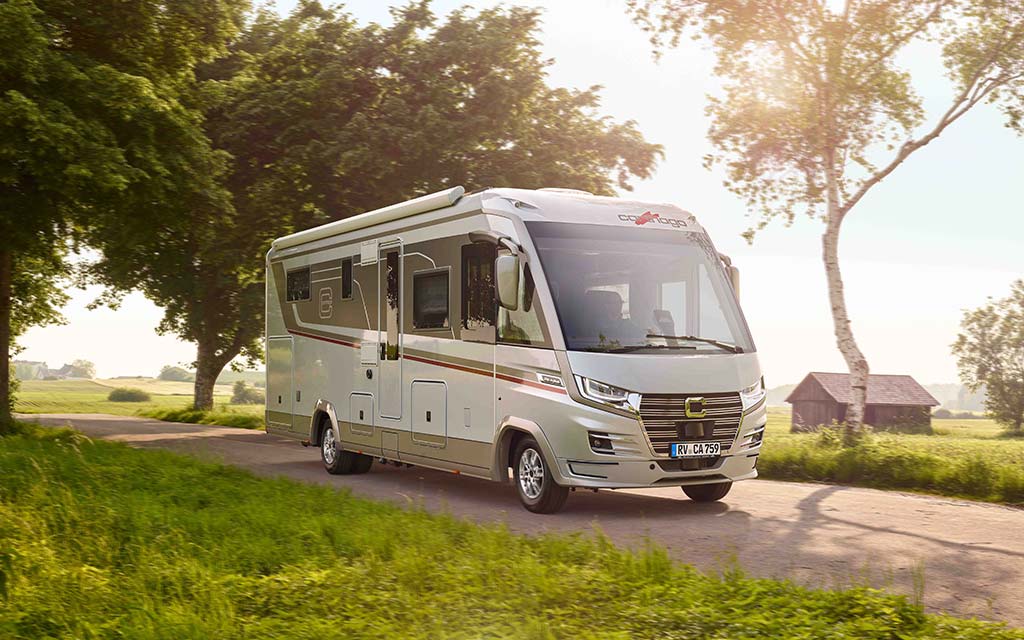 Ein moderner weißer Wohnwagen fährt auf einer sonnigen Landstraße an Bäumen vorbei