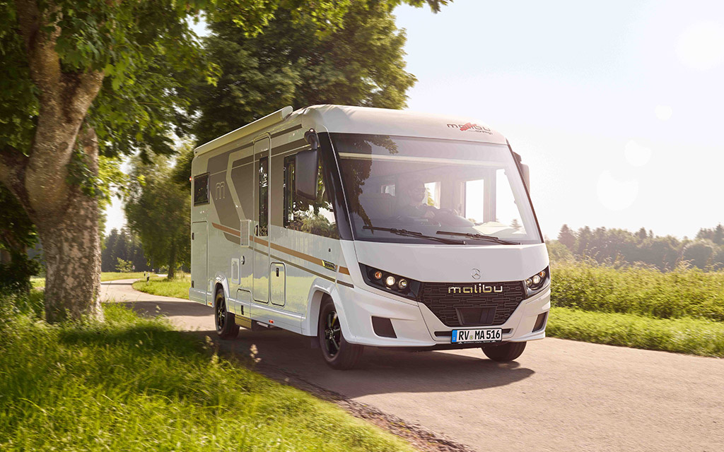 Weißes Wohnmobil fährt auf einer sonnigen Landstraße mit Bäumen und Wiesen