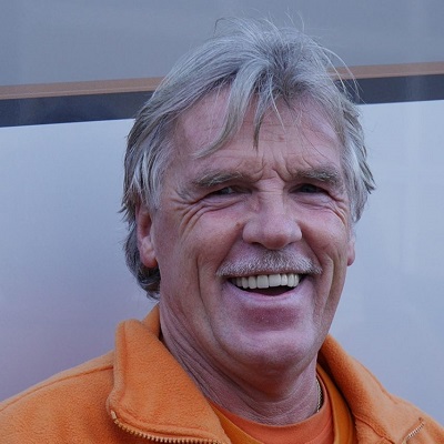 Ein älterer Mann mit grauem Haar und Schnurrbart lacht freundlich in orangener Jacke