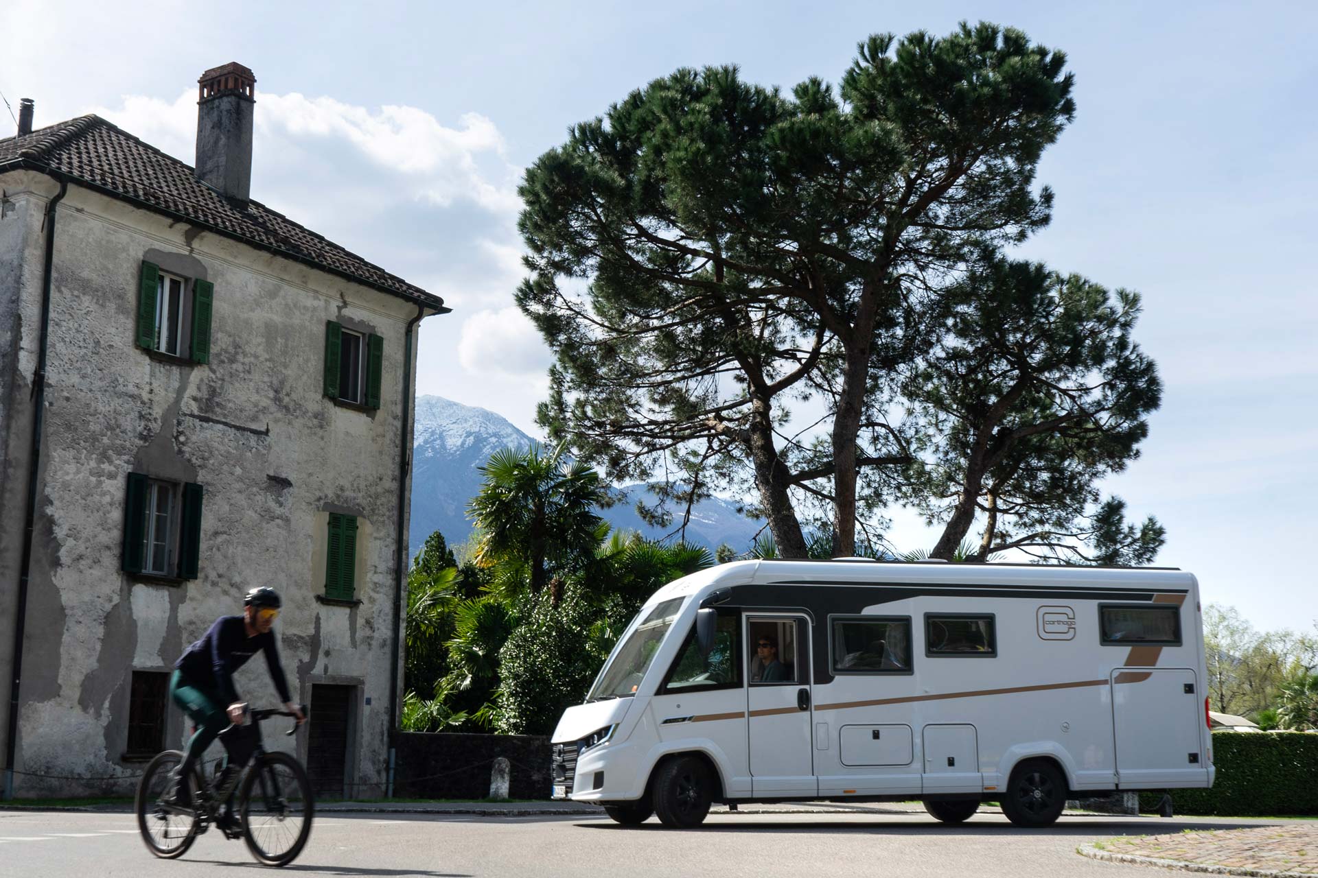 Eine Person mit Helm fährt Fahrrad vor einem weißen Wohnmobil und altem Haus bei Sonne