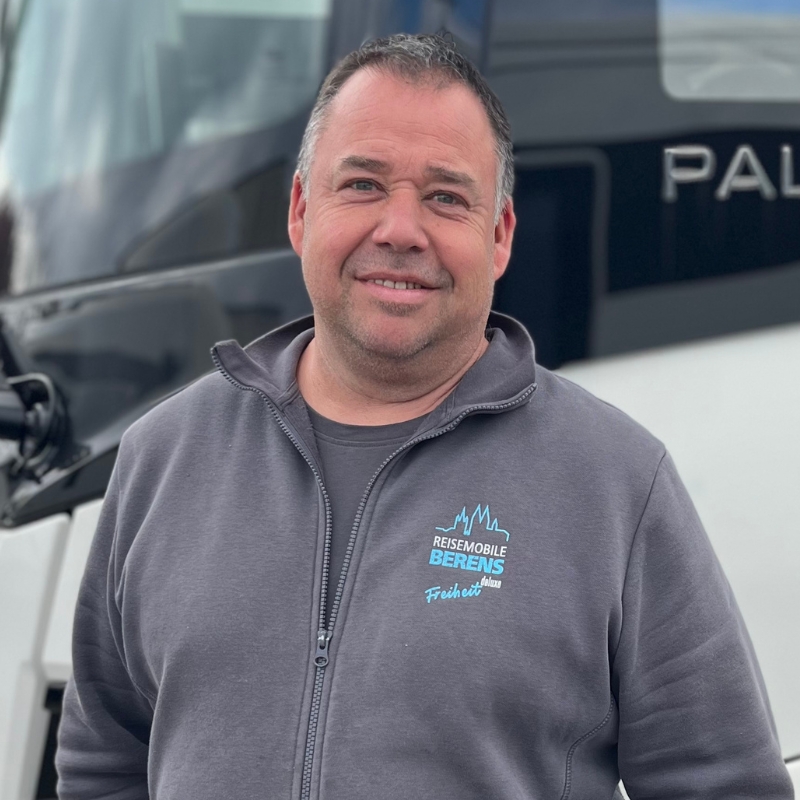 Ein lächelnder Mann mit dunklen Haaren steht vor einem weißen LKW und trägt eine graue Jacke