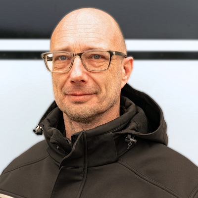 Ein älterer Mann mit Brille und Glatze lächelt freundlich in dunkler Jacke