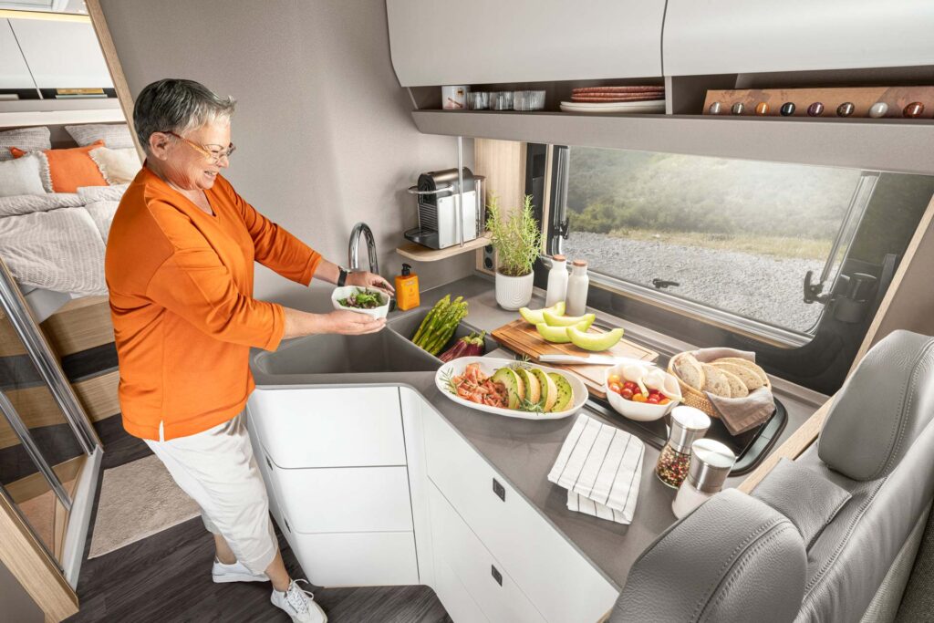 Eine ältere Frau mit kurzem grauem Haar lächelt freundlich in einer modernen Wohnmobilküche, sie trägt ein oranges Oberteil und bereitet frisches Essen zu