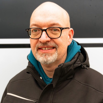Ein lächelnder älterer Mann mit Brille, Glatze und grauem Bart trägt eine schwarze Jacke