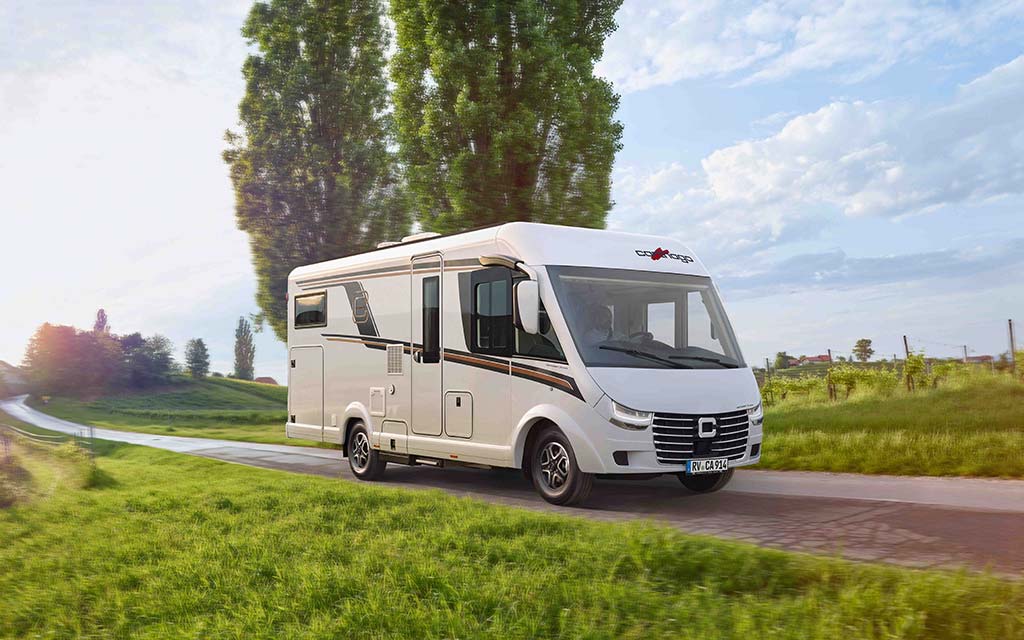 Ein weißes Wohnmobil mit schwarzen Akzenten fährt auf einer Landstraße bei Sonnenschein