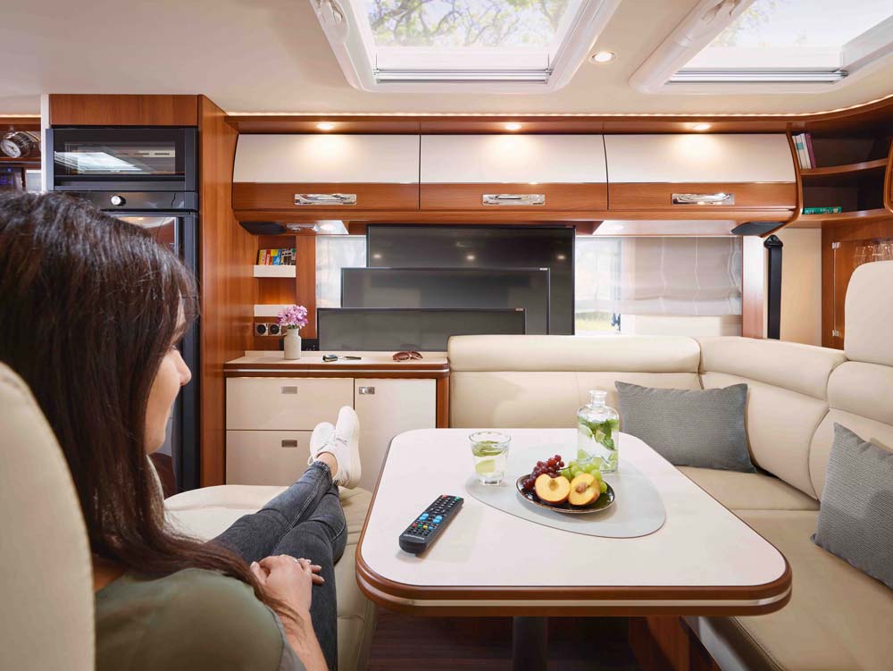 Eine Frau mit braunen Haaren sitzt entspannt auf einem beigen Sofa in einem modernen Wohnmobil mit Holzdekor und frischem Obst auf dem Tisch