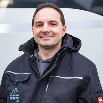 Ein lächelnder Mann mit dunklen Haaren trägt eine schwarze Jacke mit türkisfarbenem Logo