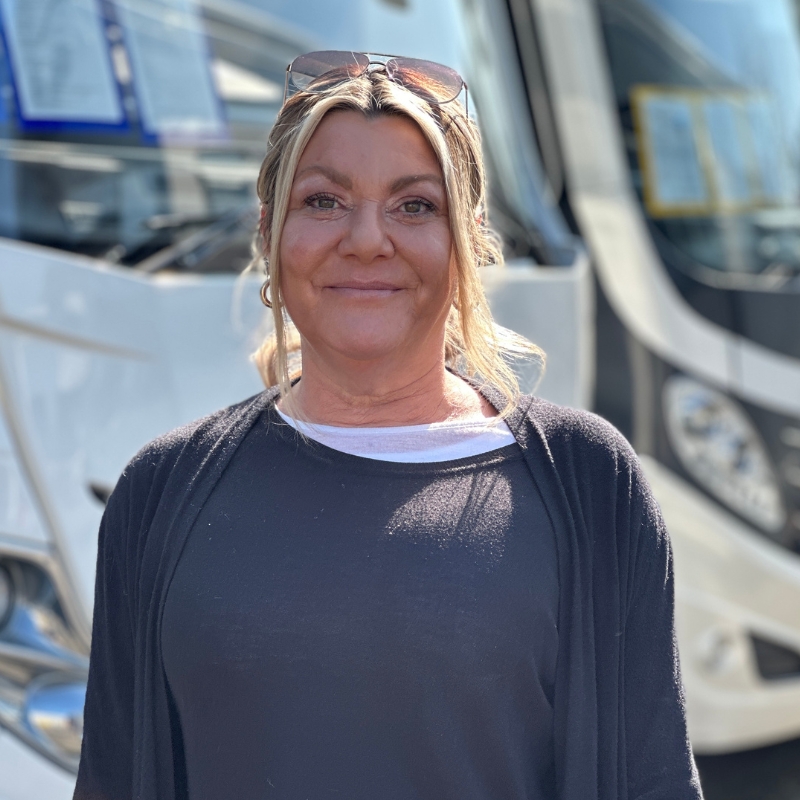 Eine lächelnde Frau mit blonden Haaren trägt eine Sonnenbrille und dunkle Kleidung
