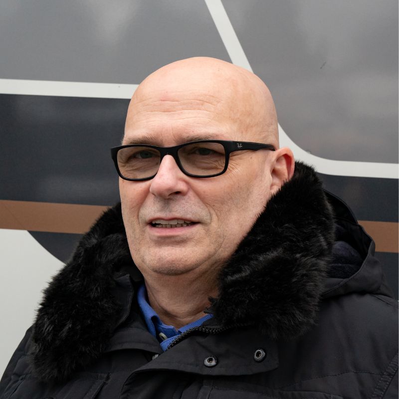 Ein älterer Mann mit Brille und Glatze lächelt leicht, trägt eine schwarze Jacke