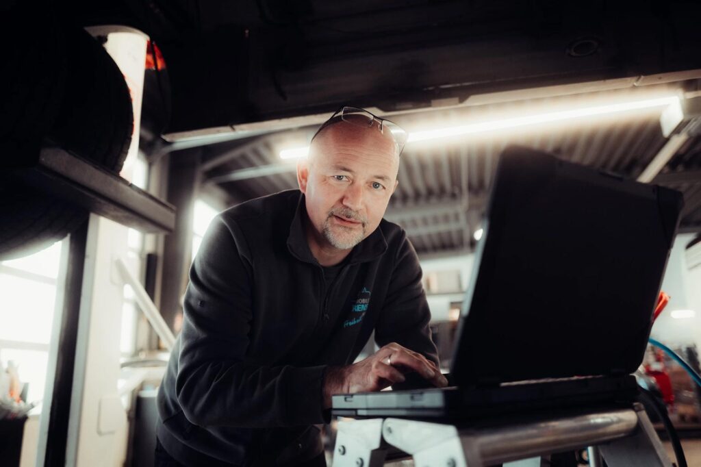 Ein älterer Mann mit Brille arbeitet konzentriert an einem schwarzen Laptop in einer Werkstatt